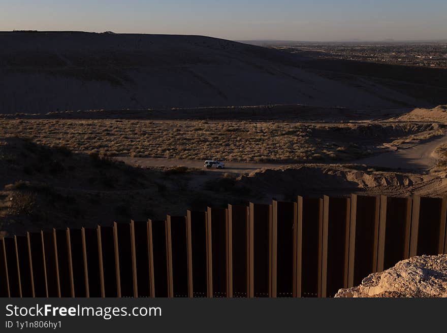 A patrol car monitors the border wall spanning a barren terrain in Ciudad Juarez at sundown, highlighting migration issues