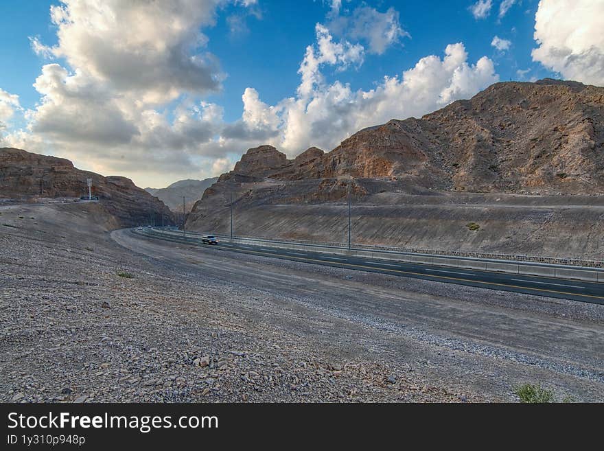Empty desert road winding through dramatic UAE mountain ranges, creating strong leading lines under clear blue skies. Limestone formations and arid valleys evoke raw natural beauty. Perfect for adventure travel campaigns, automotive ads, or metaphor visuals about life journeys