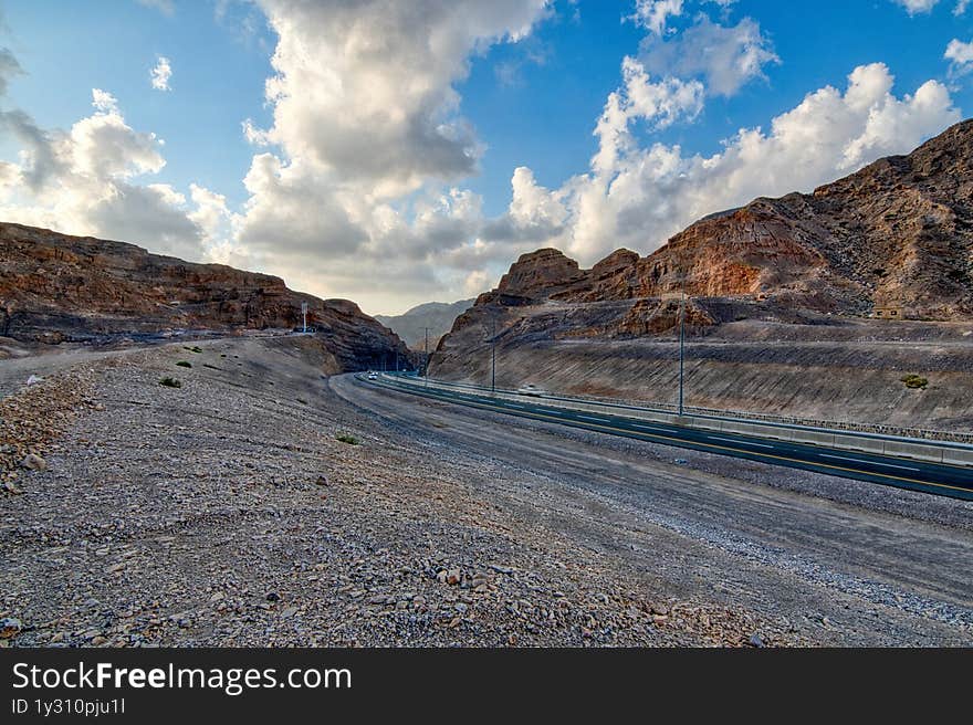 Empty desert road winding through dramatic UAE mountain ranges, creating strong leading lines under clear blue skies. Limestone formations and arid valleys evoke raw natural beauty. Perfect for adventure travel campaigns, automotive ads, or metaphor visuals about life journeys