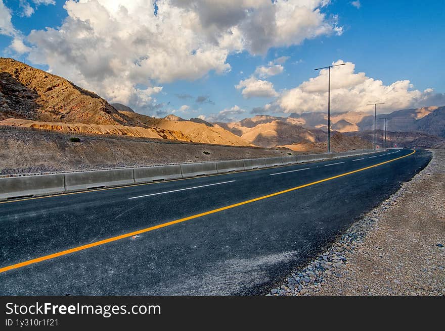Empty desert road winding through dramatic UAE mountain ranges, creating strong leading lines under clear blue skies. Limestone formations and arid valleys evoke raw natural beauty. Perfect for adventure travel campaigns, automotive ads, or metaphor visuals about life journeys