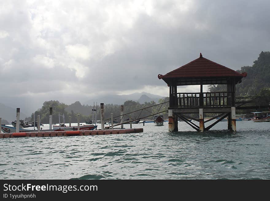 A lone fisherman s hut stands proudly on wooden stilts, its red-tiled roof a warm spot of color against the serene, gray sky. The
