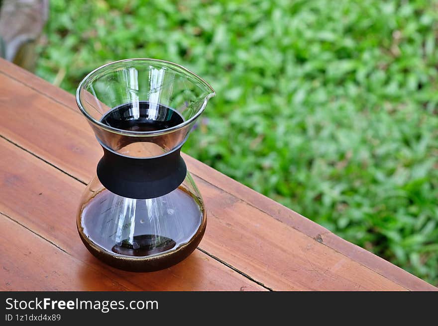 Glass coffee carafe filled with black coffee placed on wooden table with green garden background, symbolizing relaxation, morning