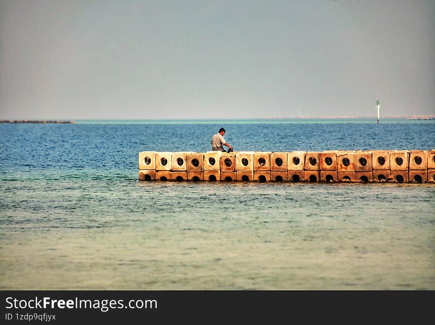 A man standing alone at the wave breaker seems to be fixing bait to catch some fish.