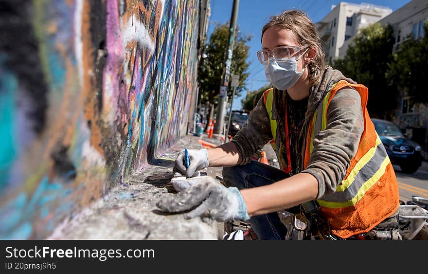 A young artist wearing safety glasses, a face mask, and a high-visibility vest carefully restores a vibrant mural on an outdoor wall. The artist is kneeling and using a small tool to clean or repair the artwork. The scene conveys themes of community, art preservation, urban renewal, and the dedication of artists to maintain public art spaces. A young artist wearing safety glasses, a face mask, and a high-visibility vest carefully restores a vibrant mural on an outdoor wall. The artist is kneeling and using a small tool to clean or repair the artwork. The scene conveys themes of community, art preservation, urban renewal, and the dedication of artists to maintain public art spaces.