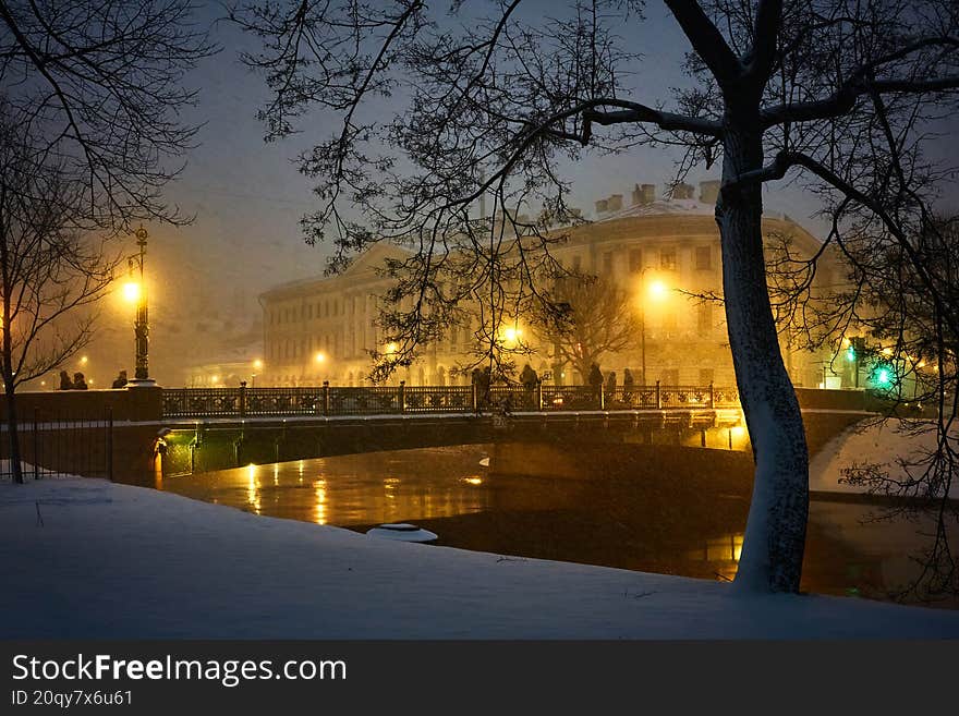 A winter night in the city. A bridge over a canal and a building in the background, illuminated by streetlights and covered in sno