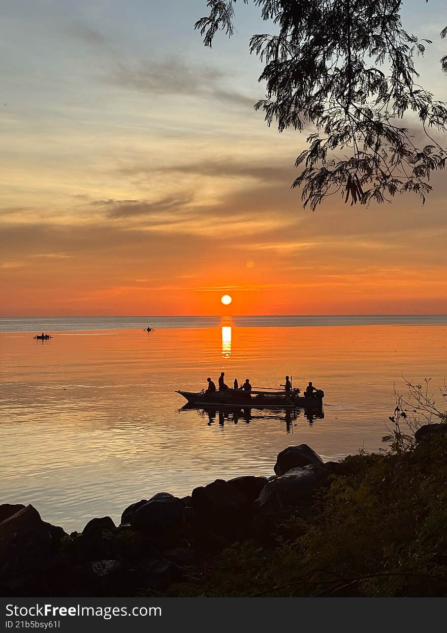 A beautiful sunset view over a calm sea. Several boats are seen as silhouettes on the water, which reflects golden orange colors, with rocks and a tree silhouette in the foreground.