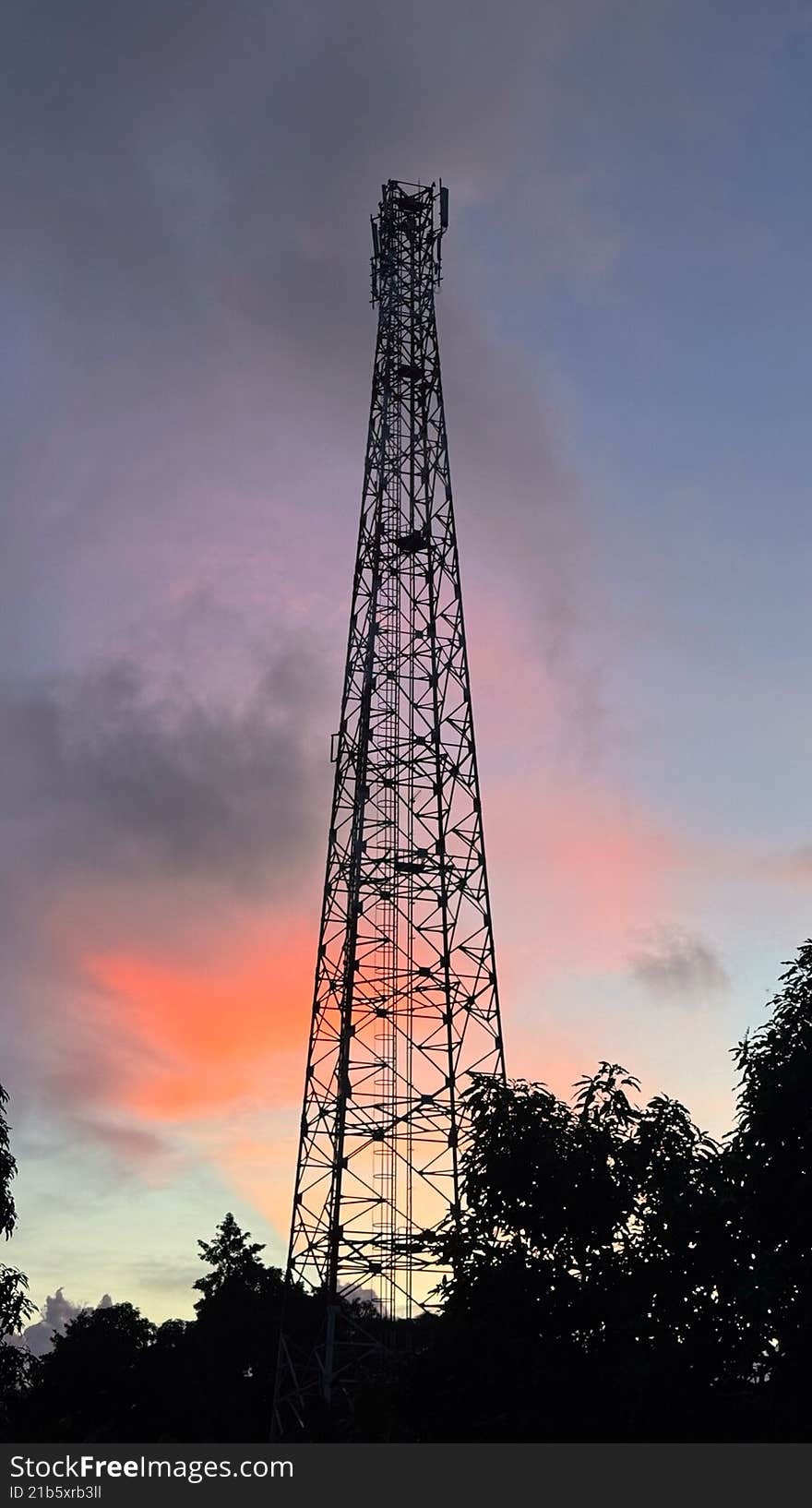 A striking silhouette of a tall telecommunication tower against a vibrant sunset sky. The sky displays hues of purple, pink, and orange, with dark foliage in the foreground completing the scene.