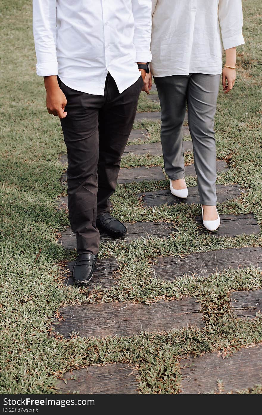 A waist-down shot of a man and woman walking together on a rustic wooden path embedded in green grass, dressed in casual smart attire