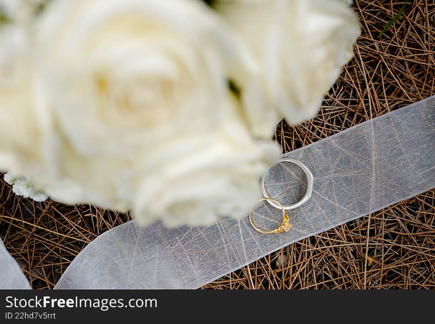 Top-down view of wedding rings on a white ribbon over dry pine needles, with a soft-focus white rose bouquet in the foreground.