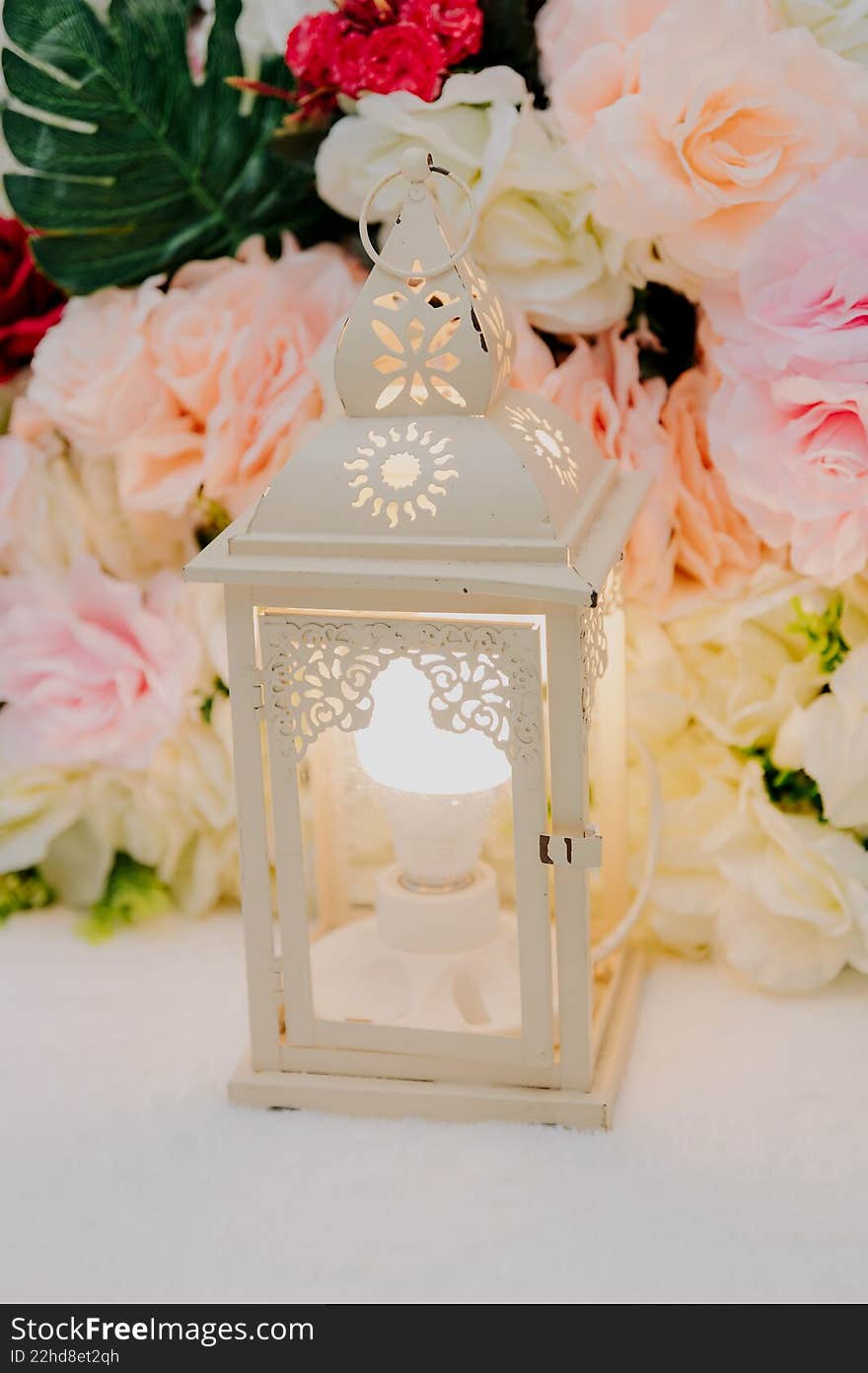 An ornamental white Moroccan-style lantern lit from within, set against a backdrop of pastel pink and white roses