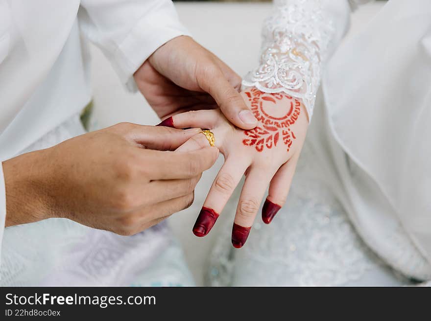 Close-up of a groom's hands sliding a gold ring onto the bride's finger, showcasing her white lace dress and red henna. Close-up of a groom's hands sliding a gold ring onto the bride's finger, showcasing her white lace dress and red henna