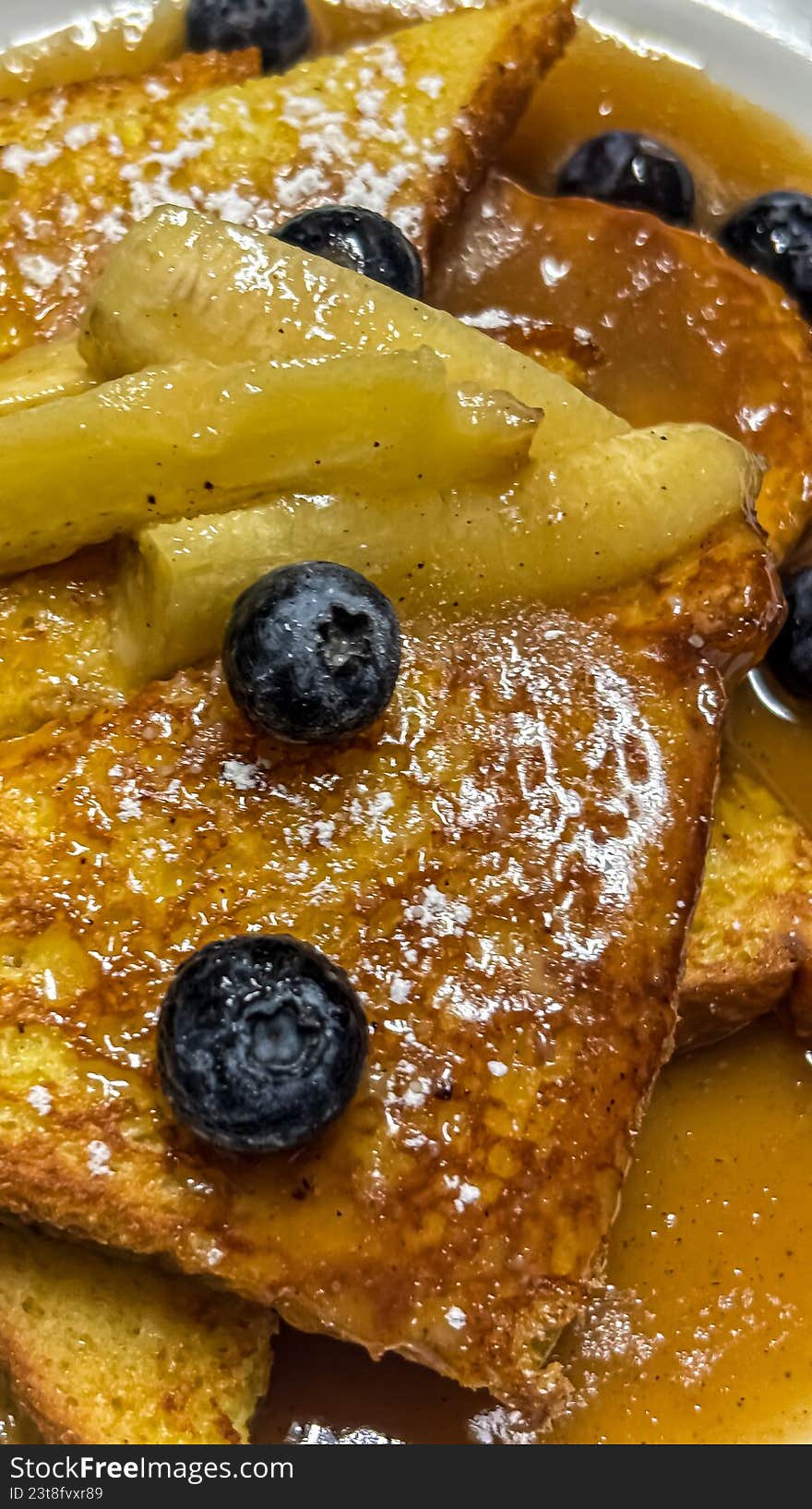 A luscious, close-up shot of thick-cut French toast triangles drizzled with warm maple syrup or caramel sauce. Topped with sautéed banana slices and vibrant fresh blueberries, this decadent breakfast dish is captured in rich detail. Perfect for brunch menus, cafe advertising, culinary blogs, or morning-themed lifestyle content.