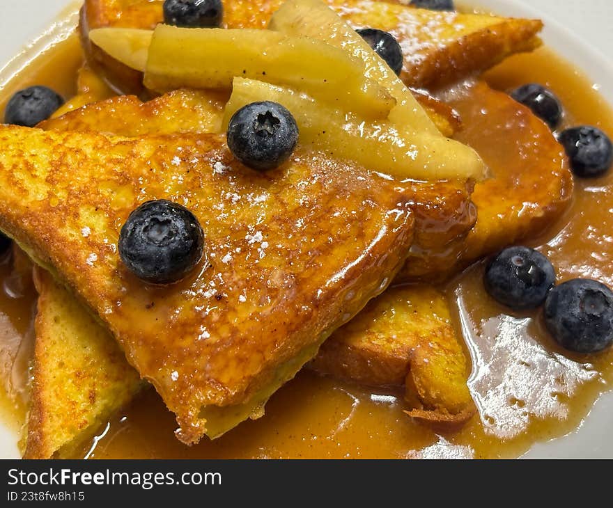 A luscious, close-up shot of thick-cut French toast triangles drizzled with warm maple syrup or caramel sauce. Topped with sautéed banana slices and vibrant fresh blueberries, this decadent breakfast dish is captured in rich detail. Perfect for brunch menus, cafe advertising, culinary blogs, or morning-themed lifestyle content.