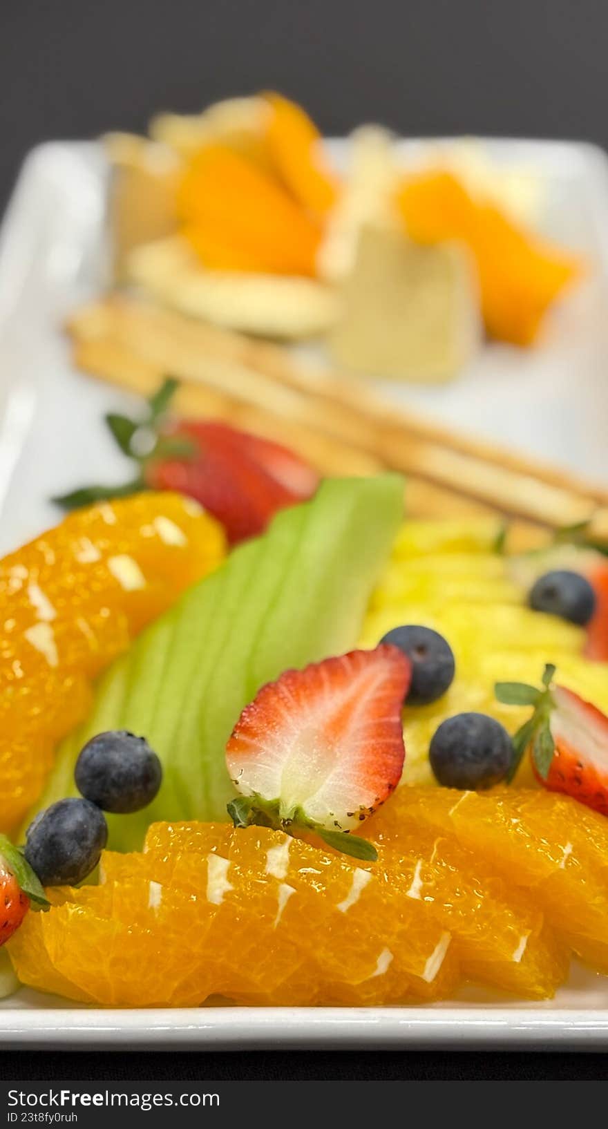 A sophisticated, close-up shot of a beautifully arranged appetizer platter. The image features a selection of fine cheeses, including creamy brie and sharp cheddar, alongside fresh strawberries, blueberries, citrus segments, and crisp breadsticks. This elegant composition is perfect for luxury catering marketing, wine pairing guides, hospitality brochures, or high-end food editorial content.