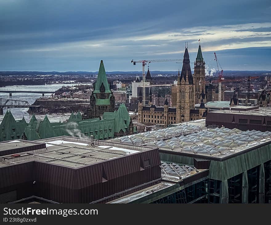A sweeping, high-angle landscape of Parliament Hill in Ottawa, Ontario, under a deep blue, overcast sky. This wide-angle shot highlights the gothic revival architecture of the Canadian Parliament buildings, the frozen Ottawa River, and active construction cranes engaged in the Centre Block restoration project. The cool color palette and urban perspective make this ideal for travel editorials, news backgrounds, and architectural publications.