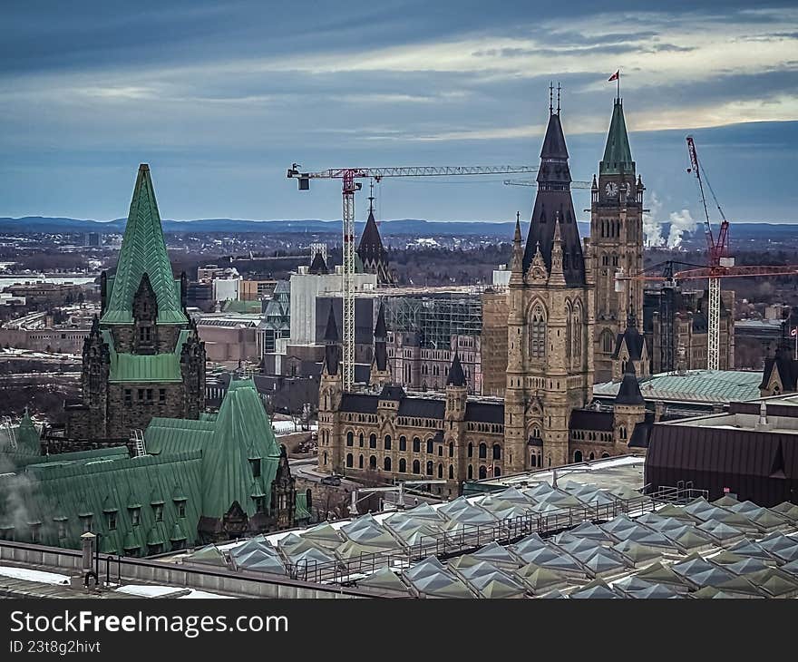 A sweeping, high-angle landscape of Parliament Hill in Ottawa, Ontario, under a deep blue, overcast sky. This wide-angle shot highlights the gothic revival architecture of the Canadian Parliament buildings, the frozen Ottawa River, and active construction cranes engaged in the Centre Block restoration project. The cool color palette and urban perspective make this ideal for travel editorials, news backgrounds, and architectural publications.