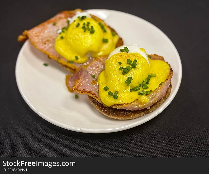 A high-detail, overhead close-up of two perfectly poached eggs served on toasted English muffins with Canadian bacon. Drizzled with a rich, velvety yellow hollandaise sauce and garnished with freshly chopped green chives, this image captures the ultimate brunch indulgence. Ideal for restaurant menus, breakfast cafe marketing, cookbooks, and food editorial content.