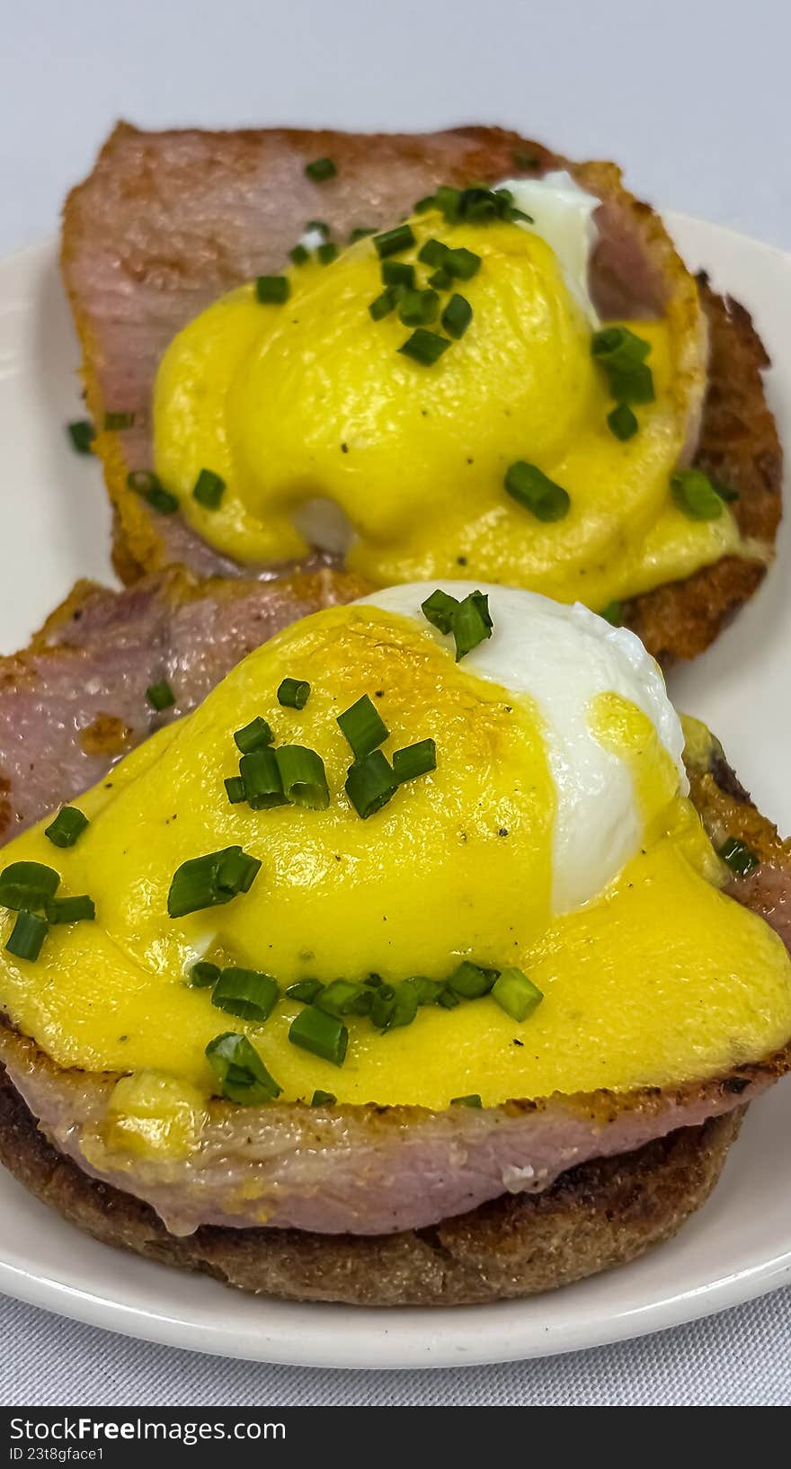 A high-detail, overhead close-up of two perfectly poached eggs served on toasted English muffins with Canadian bacon. Drizzled with a rich, velvety yellow hollandaise sauce and garnished with freshly chopped green chives, this image captures the ultimate brunch indulgence. Ideal for restaurant menus, breakfast cafe marketing, cookbooks, and food editorial content.