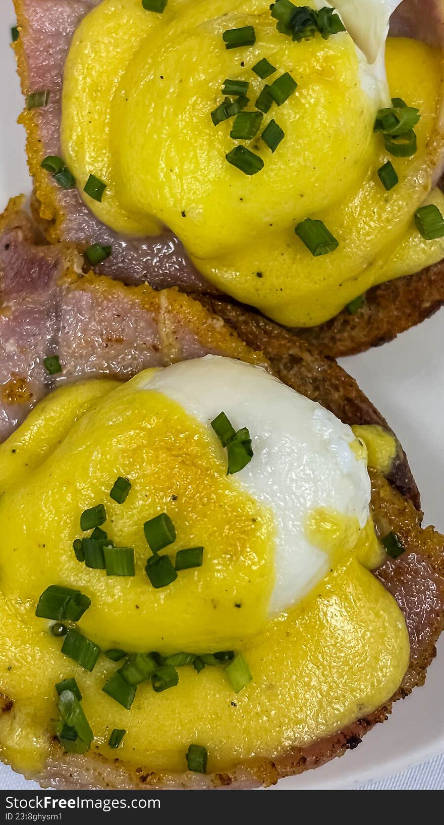A high-detail, overhead close-up of two perfectly poached eggs served on toasted English muffins with Canadian bacon. Drizzled with a rich, velvety yellow hollandaise sauce and garnished with freshly chopped green chives, this image captures the ultimate brunch indulgence. Ideal for restaurant menus, breakfast cafe marketing, cookbooks, and food editorial content.