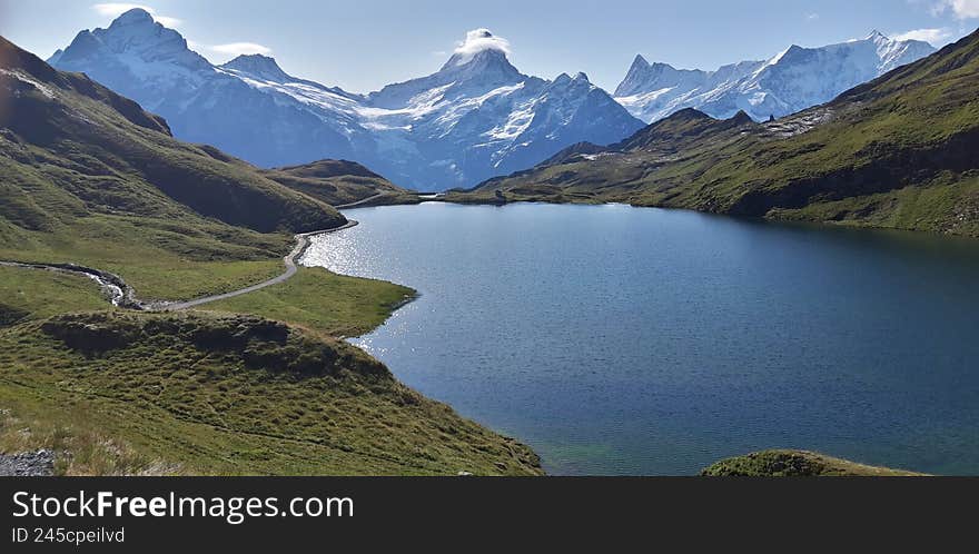Mountain landscape with a calm alpine lake surrounded by green hills and snow-capped peaks under a clear sky. Peaceful natural scenery, untouched wilderness.