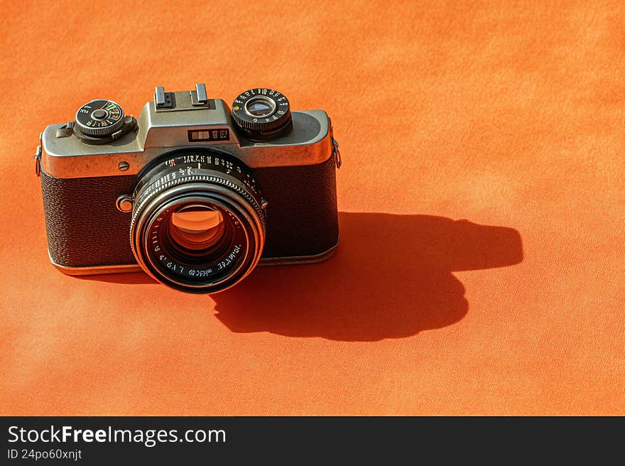 This image captures a vintage camera resting on a rustic orange background. The camera, with its metallic body and classic design, exudes a sense of nostalgia. It features a prominent lens with intricate markings, a viewfinder, and several dials and knobs indicative of its manual operation. The camera's shadow is cast on the background, adding depth to the composition. The overall scene highlights. This image captures a vintage camera resting on a rustic orange background. The camera, with its metallic body and classic design, exudes a sense of nostalgia. It features a prominent lens with intricate markings, a viewfinder, and several dials and knobs indicative of its manual operation. The camera's shadow is cast on the background, adding depth to the composition. The overall scene highlights