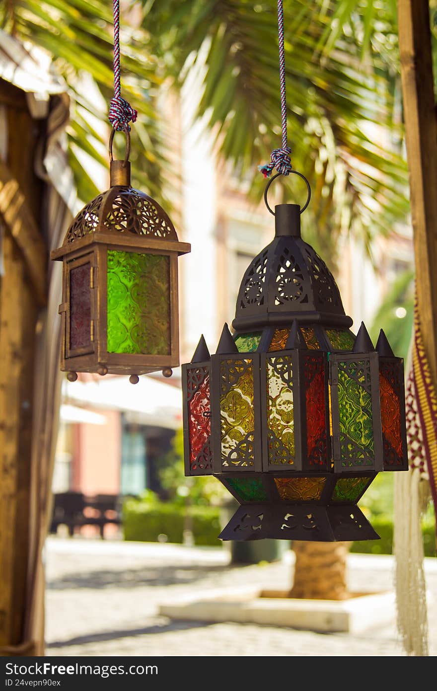 Two traditional Moroccan glass lanterns hang gracefully against the backdrop of a sunlit palm tree. The warm light highlights the delicate metalwork and colorful glass panels, emphasizing the craftsmanship and ornate details typical of Moroccan design. The soft natural atmosphere, combined with tropical greenery in the background, creates a serene and exotic ambiance. This image captures the fusion of cultural artistry and outdoor tranquility, making it ideal for travel visuals, decor themes, lifestyle imagery, or editorial content focused on Moroccan style and Mediterranean aesthetics. Two traditional Moroccan glass lanterns hang gracefully against the backdrop of a sunlit palm tree. The warm light highlights the delicate metalwork and colorful glass panels, emphasizing the craftsmanship and ornate details typical of Moroccan design. The soft natural atmosphere, combined with tropical greenery in the background, creates a serene and exotic ambiance. This image captures the fusion of cultural artistry and outdoor tranquility, making it ideal for travel visuals, decor themes, lifestyle imagery, or editorial content focused on Moroccan style and Mediterranean aesthetics.