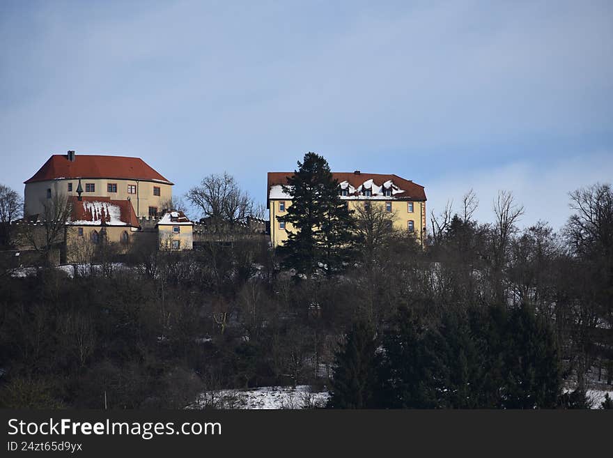 Hilltop view of Reichelsheim, Germany, featuring a historic yellow manor with red-tiled roofs, partially covered in snow. Surrounded by leafless trees and evergreen conifers, the buildings stand prominently against the clear blue sky. The architecture reflects traditional German style, and the snow adds a serene, wintry feel to the scene. The landscape blends the natural and built environment harmoniously.