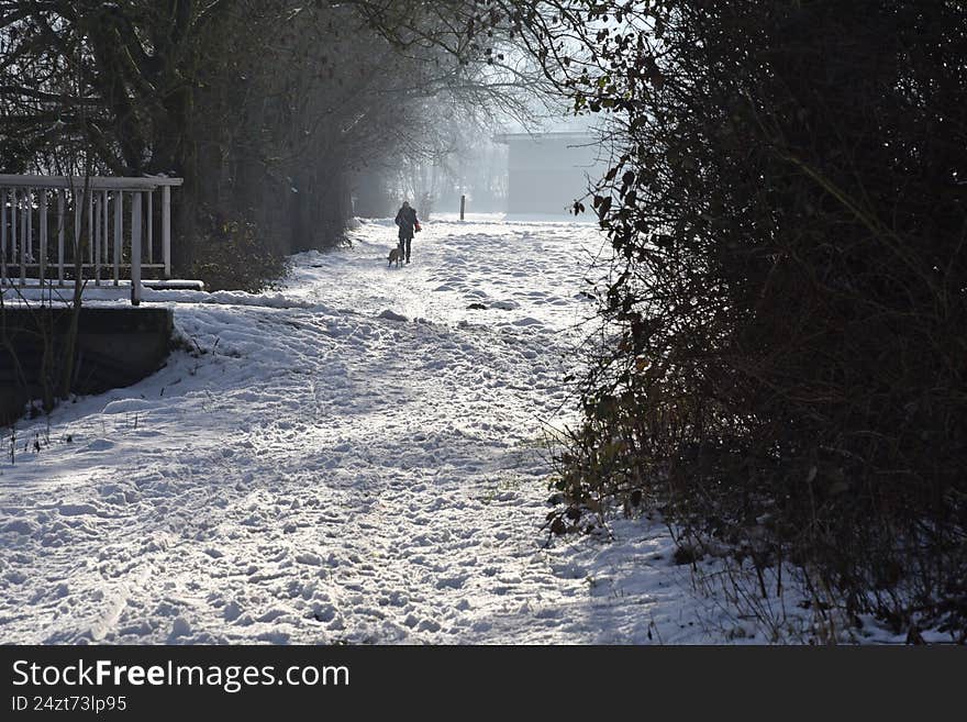Snow-covered riverside promenade in a tranquil setting. A person walks a dog along the path, surrounded by trees and dense shrubs. The ground is blanketed with snow, and a structure is visible in the distance, partially obscured by mist. Soft sunlight filters through the trees, casting gentle shadows on the snow. The scene conveys a peaceful, wintry atmosphere.