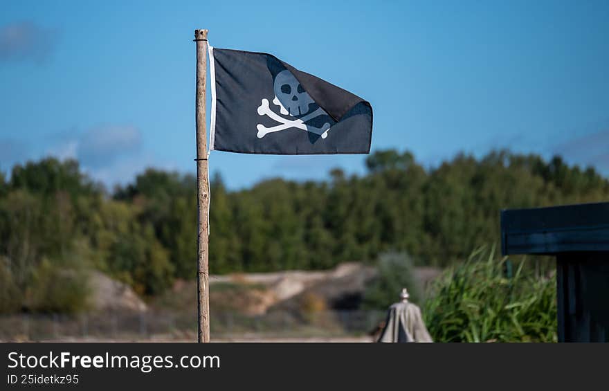 Jolly roger flag waving on a wooden pole against a clear blue sky and green trees
