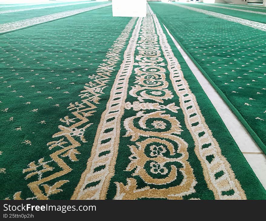 Indonesia. A low-angle perspective view of green mosque prayer carpets arranged in neat parallel rows inside a large prayer hall. The carpet features intricate beige and cream Islamic geometric and floral patterns running vertically toward the mihrab area, creating a strong sense of depth and symmetry. The soft texture and repeating motifs highlight the serene atmosphere of the sacred space. The bright natural lighting enhances the vivid green tones, emphasizing cleanliness and order within the mosque interior. No people are visible, focusing attention on the architectural and spiritual ambiance of the Islamic worship environment.